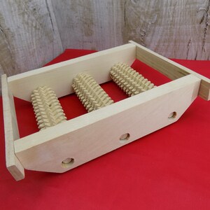 May include: Wooden foot massage roller with three rollers. The rollers are made of wood and have a textured surface. The foot massage roller is on a red surface.