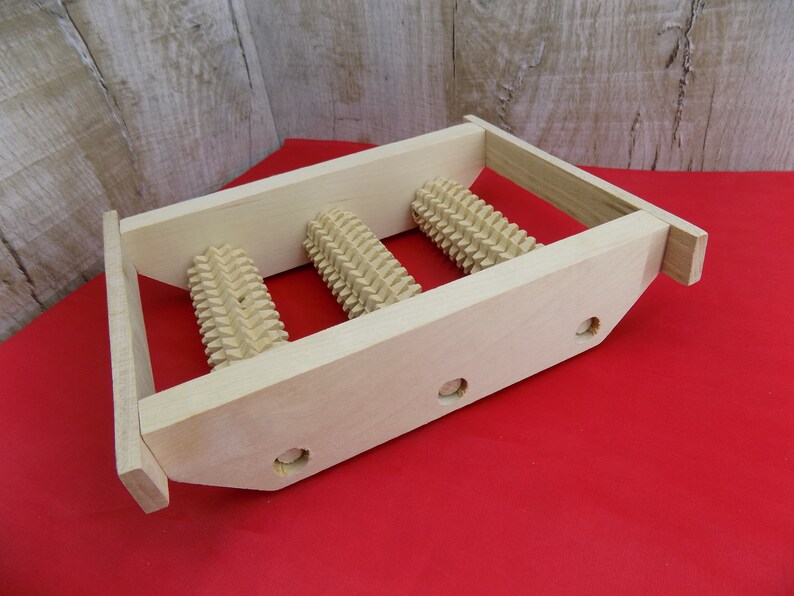 May include: Wooden foot massage roller with three rollers. The rollers are made of wood and have a textured surface. The foot massage roller is on a red surface.