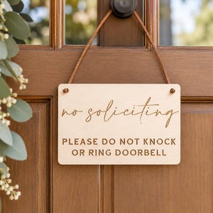 May include: A rectangular light-colored wooden sign hanging by a thin brown leather strap. The sign features the text no soliciting in a cursive script font, followed by the text PLEASE DO NOT KNOCK OR RING DOORBELL in a block capital font.