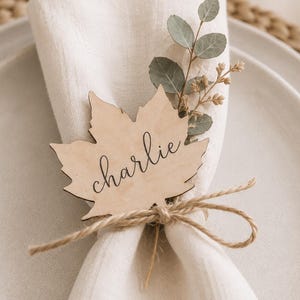 May include: A light-colored wooden place card shaped like a maple leaf with the name charlie written in black cursive script. The leaf is tied to a folded white fabric napkin with a piece of brown twine and decorated with a sprig of green eucalyptus.