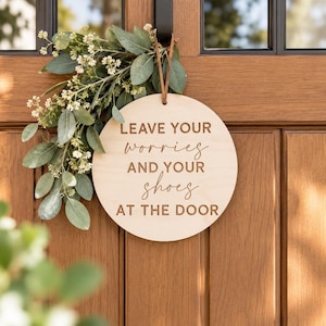 May include: A circular light-colored wooden sign hanging by a brown leather strap, featuring the engraved text LEAVE YOUR worries AND YOUR shoes AT THE DOOR, accented with a decorative sprig of green eucalyptus leaves and small white flowers.
