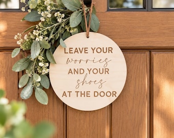 May include: A circular light-colored wooden sign hanging by a brown leather strap, featuring the engraved text LEAVE YOUR worries AND YOUR shoes AT THE DOOR, accented with a decorative sprig of green eucalyptus leaves and small white flowers.