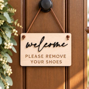 May include: A rectangular light-colored wooden sign hanging from a brown leather strap. The sign features the word welcome in black script lettering above the text PLEASE REMOVE YOUR SHOES in a simple, dark, capitalized sans-serif font.