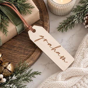 May include: A rectangular light-colored wooden gift tag with a hole at the top, featuring the name "graham" engraved in a cursive script. The tag is attached to a brown paper-wrapped gift by a thin, flat, light brown leather cord.