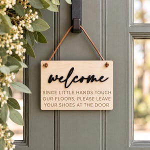 May include: A rectangular light-colored wooden sign hanging from a brown leather strap. The sign features the black 3D script text "welcome" above the engraved text "SINCE LITTLE HANDS TOUCH OUR FLOORS, PLEASE LEAVE YOUR SHOES AT THE DOOR."