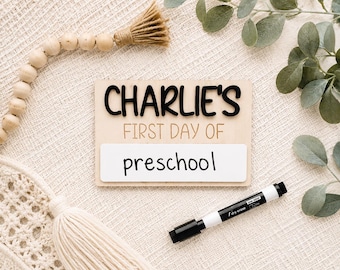 May include: A rectangular wooden sign with the black 3D text CHARLIE'S and the brown text FIRST DAY OF printed above a white dry-erase section with the word preschool written in black marker, accompanied by a black dry-erase marker.