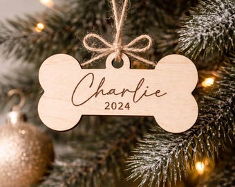 May include: A light-colored wooden ornament shaped like a dog bone, featuring the name Charlie and the year 2024 engraved in black script and block text. The ornament is suspended by a thin, natural-colored jute twine tied into a small bow.
