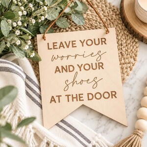 May include: A light-colored wooden sign with a pennant-shaped bottom edge, featuring the engraved text LEAVE YOUR worries AND YOUR shoes AT THE DOOR. The sign is suspended by a thin brown leather cord attached to the top corners of the wood.