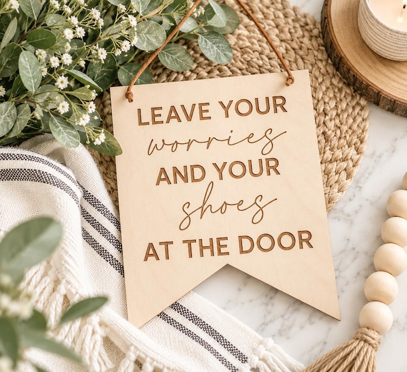 May include: A light-colored wooden sign with a pennant-shaped bottom edge, featuring the engraved text LEAVE YOUR worries AND YOUR shoes AT THE DOOR. The sign is suspended by a thin brown leather cord attached to the top corners of the wood.