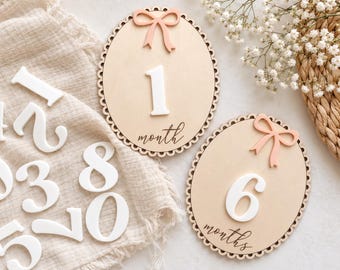 May include: Two oval wooden plaques with scalloped edges, each featuring a peach-colored bow at the top and a large white number. One plaque displays the number 1 and the word month, while the other shows the number 6 and the word months.
