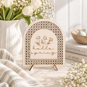 May include: An arched wooden sign featuring a light wood finish and a woven cane pattern border. The center displays a laser-engraved illustration of three wildflowers above the cursive text "hello spring" in a dark brown ink.