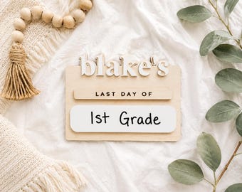 May include: A rectangular wooden sign featuring the white 3D text blake's at the top, a horizontal wooden strip with the black text LAST DAY OF, and a white rectangular insert with the black handwritten-style text 1st Grade printed on it.