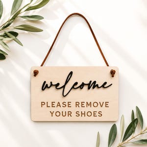 May include: A rectangular light-colored wooden sign with a brown leather hanging strap. The sign features the black script text "welcome" above the block-lettered text "PLEASE REMOVE YOUR SHOES" in a dark brown color, with rounded corners.