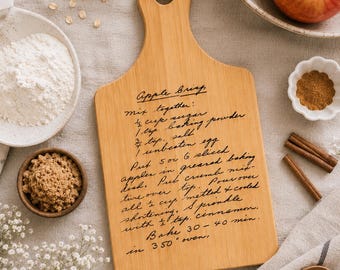 May include: A light-colored wooden cutting board with a handle and a hole for hanging, featuring a handwritten recipe for Apple Crisp in black ink. The text includes instructions for mixing sugar, baking powder, salt, an egg, apples, and cinnamon.