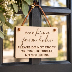 May include: A rectangular light-colored wooden sign with a leather strap for hanging. The sign features the text "working from home" in a cursive script, followed by "PLEASE DO NOT KNOCK OR RING DOORBELL. NO SOLICITING" in a block font.