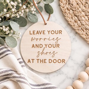 May include: A round, light-colored wooden sign featuring the engraved text "LEAVE YOUR worries AND YOUR shoes AT THE DOOR" in a combination of block and script fonts. The sign includes a thin brown leather cord for hanging at the top center.