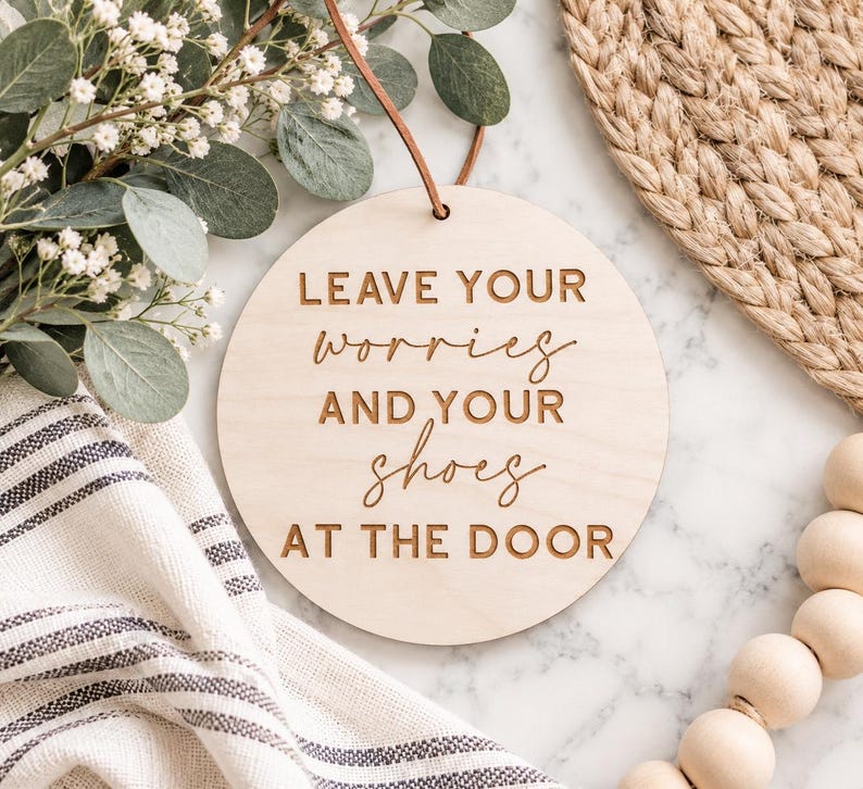 May include: A round, light-colored wooden sign featuring the engraved text "LEAVE YOUR worries AND YOUR shoes AT THE DOOR" in a combination of block and script fonts. The sign includes a thin brown leather cord for hanging at the top center.