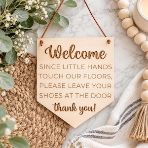 May include: A light-colored wooden sign with a pointed bottom edge, featuring the engraved text: Welcome, SINCE LITTLE HANDS TOUCH OUR FLOORS, PLEASE LEAVE YOUR SHOES AT THE DOOR thank you! The sign hangs from a thin brown leather cord.