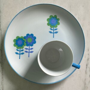 Puede incluir: Un plato de cer&aacute;mica blanco con un borde azul y un dise&ntilde;o floral azul y verde. Una taza de cer&aacute;mica blanca con un asa azul se encuentra sobre el plato.