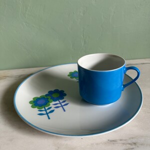 Puede incluir: Un plato de cer&aacute;mica blanco con un borde azul y un dise&ntilde;o floral en tonos azules, verdes y morados. Una taza de cer&aacute;mica azul con interior blanco se encuentra sobre el plato.