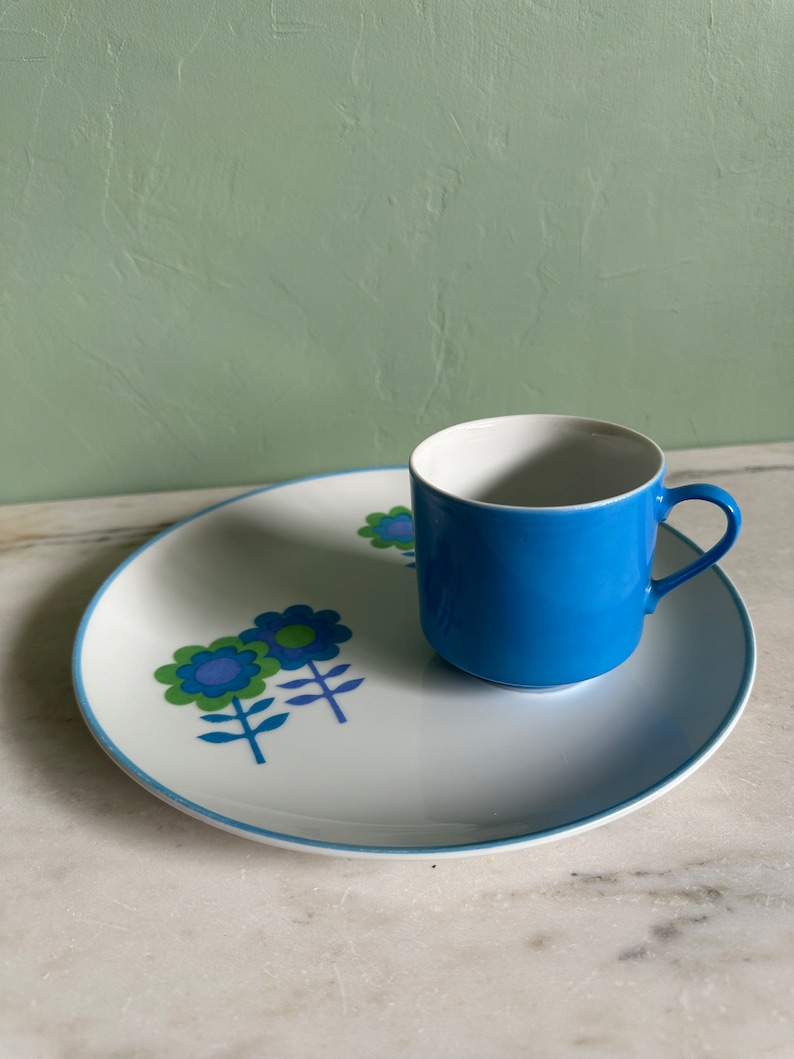 Puede incluir: Un plato de cer&aacute;mica blanco con un borde azul y un dise&ntilde;o floral en tonos azules, verdes y morados. Una taza de cer&aacute;mica azul con interior blanco se encuentra sobre el plato.