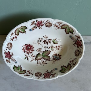 May include: A white oval serving bowl with a brown and green floral pattern. The bowl has a scalloped edge and is decorated with small red, brown, and blue flowers.