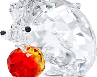 Puede incluir: Una figura de erizo de cristal transparente con ojos negros y nariz negra. El erizo sostiene una manzana facetada roja y amarilla. La figura está hecha de cristal transparente y tiene un diseño geométrico.