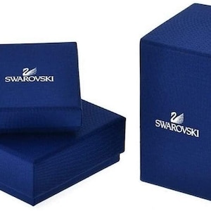 May include: Two blue boxes with the Swarovski logo. The top box is smaller and the bottom box is larger. Both boxes have a textured surface.