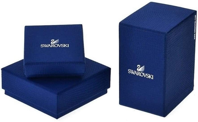 May include: Two blue boxes with the Swarovski logo. The top box is smaller and the bottom box is larger. Both boxes have a textured surface.