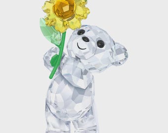 May include: A crystal bear figurine holding a yellow sunflower with a green stem and leaf. The bear has black eyes and a smiling mouth. The figurine is made of clear, faceted crystal.