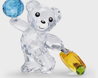 May include: A clear crystal bear figurine with a blue crystal ball held in its left hand and a yellow and green crystal object in its right hand. The bear has black eyes and a smiling face.