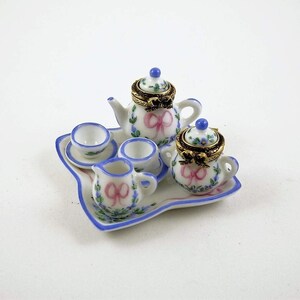 May include: Miniature porcelain tea set with a teapot, creamer, sugar bowl, and two cups on a tray. The set features a floral design with pink bows and blue accents. The teapot and sugar bowl have gold-colored rims.