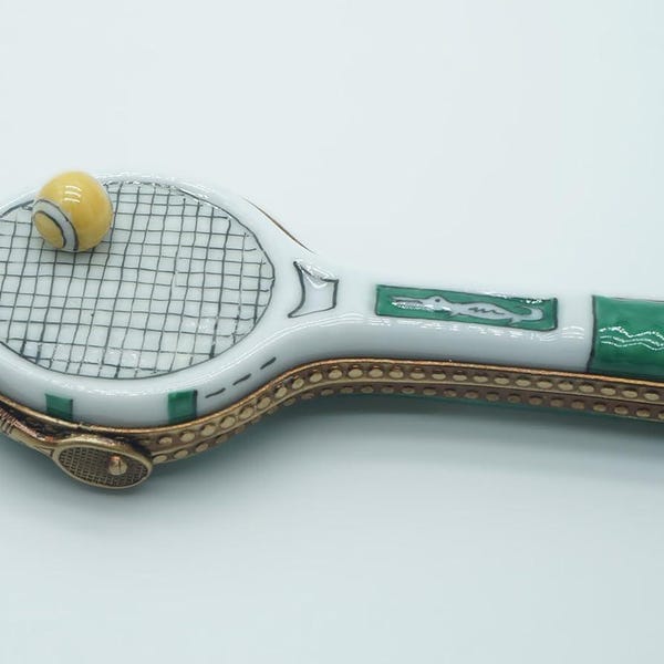 May include: A porcelain trinket box shaped like a tennis racket with a white head, a black grid pattern, and a yellow ball attached. The handle is white and green with a small alligator graphic, featuring a gold-toned metal hinge and clasp.