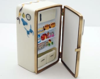 Puede incluir: Un mini refrigerador de porcelana blanca con ribete dorado y una puerta abierta. El interior del refrigerador está lleno de artículos de comida en miniatura, incluyendo leche, mantequilla, fruta y pasteles. El exterior del refrigerador tiene un reloj y un sujetador azul.