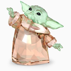 May include: A crystal figurine of a green alien character from the Star Wars franchise, known as "The Child" or "Baby Yoda", wearing a peach-colored robe. The figurine is made of clear crystal with a green tint for the character's skin and a peach tint for the robe.