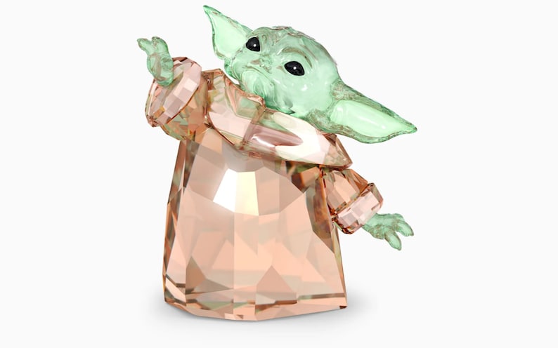 May include: A crystal figurine of a green alien character from the Star Wars franchise, known as "The Child" or "Baby Yoda", wearing a peach-colored robe. The figurine is made of clear crystal with a green tint for the character's skin and a peach tint for the robe.