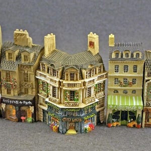 May include: A set of five miniature Parisian buildings with detailed facades, each with a unique storefront and signage. The buildings are painted in shades of beige, brown, and white, with accents of green and red. The storefronts feature signs that read "Bistro de Paris", "Antiquities de Paris", "Chez Louis", and "Le Petit Cafe".