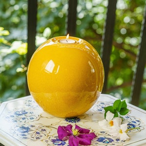 May include: A spherical, glossy, yellow candle holder with a lit candle inside. The candle holder sits on a white hexagonal table with blue floral designs. The background features greenery and a blurred view of a window.