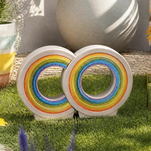 May include: Two white, circular garden ornaments with rainbow-colored inner rings. The ornaments are placed on a bed of green grass, surrounded by colorful flowers and potted plants. A candle lantern is also visible.