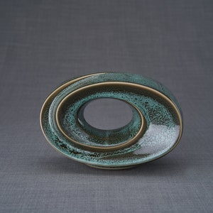 This unique keepsake urn represents a horizontal ellipse with a hole in the middle. Shaped as a spiral, a vortex, this handmade memorial urn holds the ashes in its walls. This keepsake is made of ceramic and comes in oily green melange color.