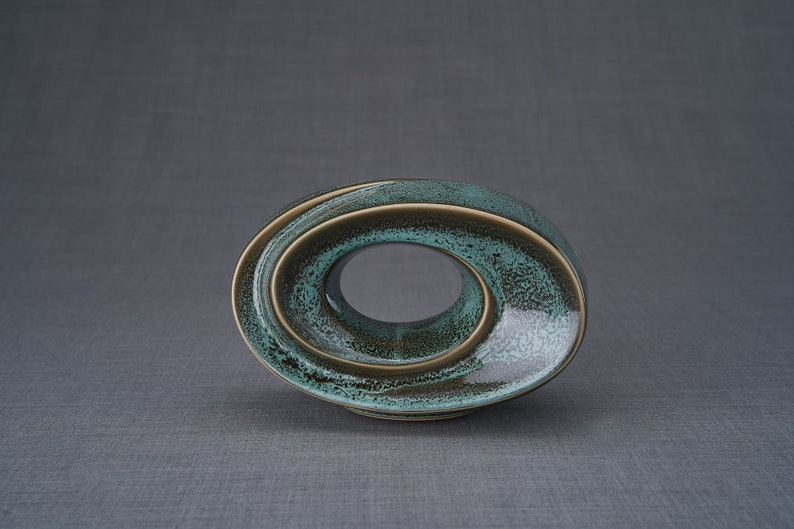 This unique keepsake urn represents a horizontal ellipse with a hole in the middle. Shaped as a spiral, a vortex, this handmade memorial urn holds the ashes in its walls. This keepsake is made of ceramic and comes in oily green melange color.