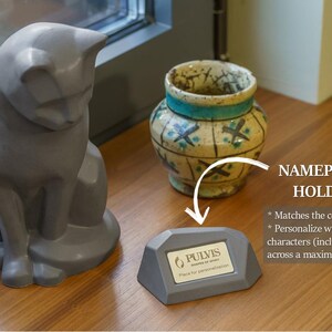 a gray ceramic cat figurine sitting on a wooden surface next to a ceramic vase. There is also a small gray plaque or holder with text on it, which appears to be a nameplate or holder.