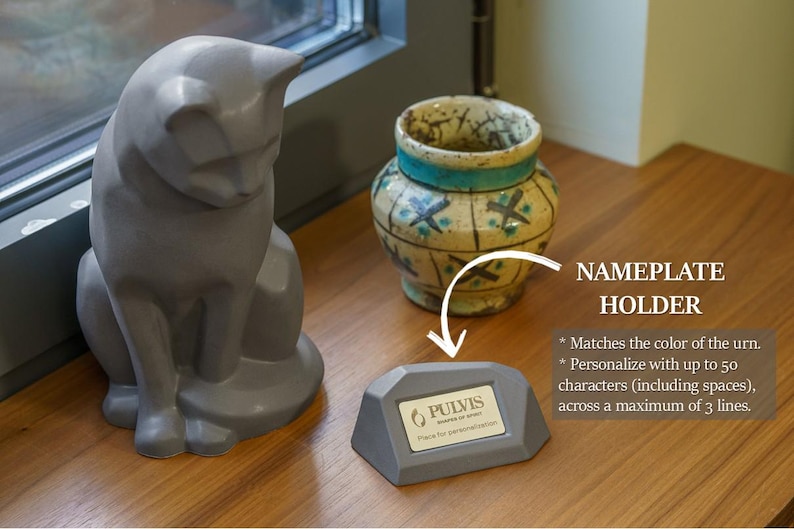 a gray ceramic cat figurine sitting on a wooden surface next to a ceramic vase. There is also a small gray plaque or holder with text on it, which appears to be a nameplate or holder.