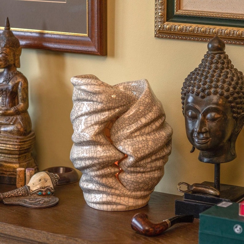 May include: A cream-colored ceramic vase with a unique, textured design. The vase has a crackle glaze and a twisted, organic shape. The interior glows with a warm light. Decorative items, including a Buddha statue and a pipe, are in the background.