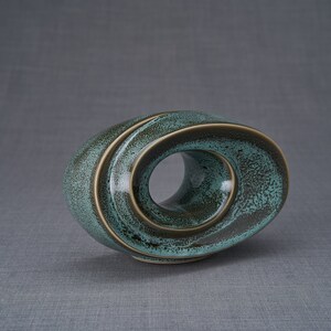 This unique keepsake urn represents a horizontal ellipse with a hole in the middle. Shaped as a spiral, a vortex, this handmade memorial urn holds the ashes in its walls. This keepsake is made of ceramic and comes in oily green melange color.