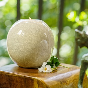 May include: A spherical, off-white ceramic candle holder with a crackle glaze finish. A small flame flickers atop the candle holder, which sits on a wooden surface. A white flower and green leaves are placed on the wooden surface.