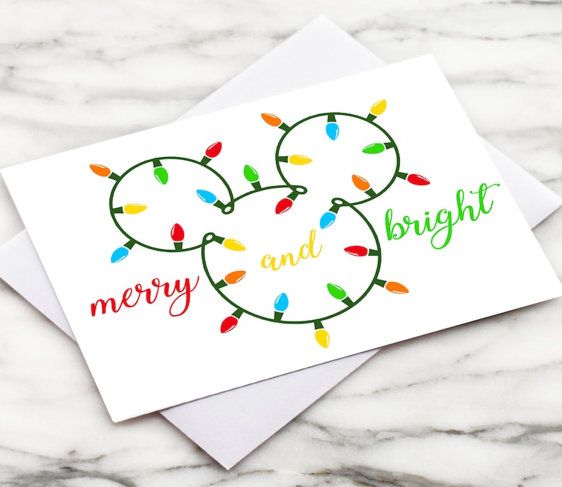 Puede incluir: Una tarjeta blanca con un contorno verde de una cabeza de Mickey Mouse hecha de luces navide&ntilde;as de colores. El texto "merry and bright" est&aacute; escrito en verde y rojo.