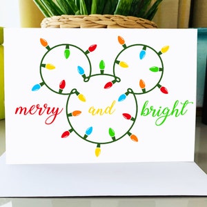 Puede incluir: Una tarjeta blanca con una cadena de luces verdes en forma de la cabeza de Mickey Mouse. Las luces son rojas, amarillas, verdes, azules y naranjas. El texto "merry and bright" est&aacute; escrito en cursiva roja y verde.