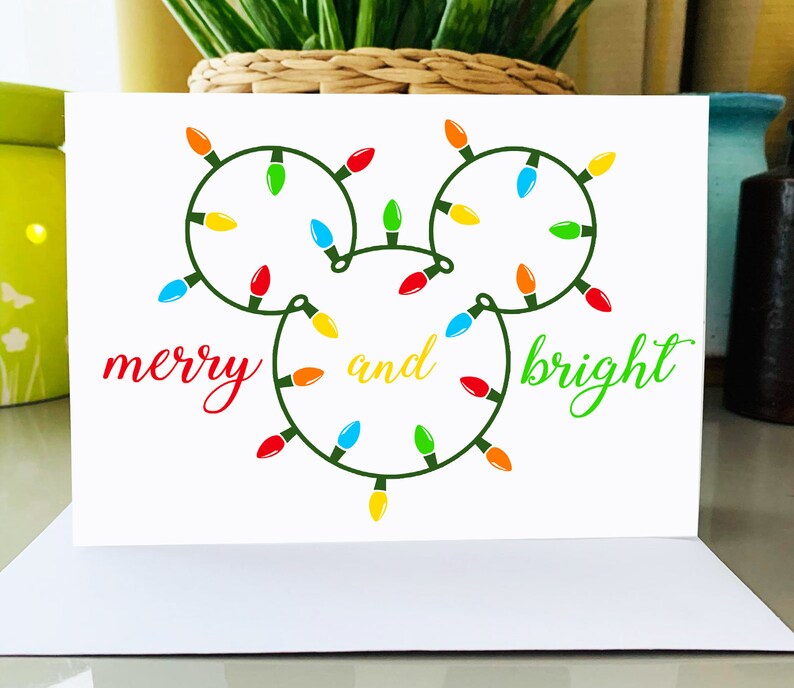 Puede incluir: Una tarjeta blanca con una cadena de luces verdes en forma de la cabeza de Mickey Mouse. Las luces son rojas, amarillas, verdes, azules y naranjas. El texto "merry and bright" est&aacute; escrito en cursiva roja y verde.