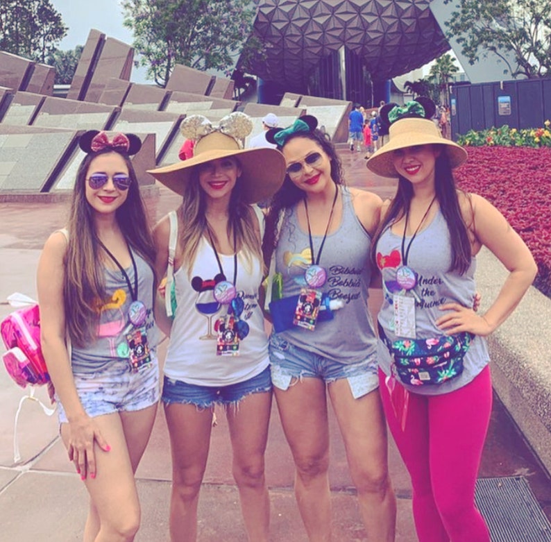 May include: Four friends wearing grey tank tops with different Disney-themed designs and mouse ears. The tank tops have the text "Drink Like a Princess", "Biddles Beebis Beest", "Under the Rum Ham", and "Mickey Mouse" on them. The friends are standing in front of a building with a large, futuristic structure in the background.